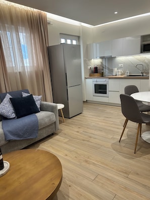 A modern kitchen with a refrigerator, oven, and sink. There is a dining table with chairs and a sofa with a throw blanket.