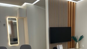 Un soggiorno moderno con un tavolo da pranzo rotondo, sedie grigie rivestite in tessuto, un tavolino in legno, una TV a schermo piatto montata su una parete rivestita in legno e un mobile TV bianco con un vaso decorativo.