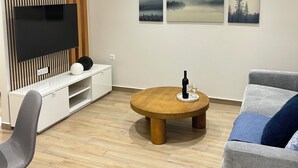 Un soggiorno moderno con una TV a schermo piatto, una parete rivestita in legno, un tavolo rotondo in legno con una bottiglia e dei bicchieri, un divano grigio e tre quadri appesi al muro.