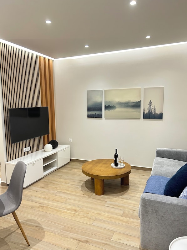 A modern living room with a flat-screen TV, wooden panel wall, a round wooden table with a bottle and glasses, a grey sofa, and three framed pictures on the wall.