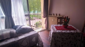 A room with two beds, a table with a red placemat, a potted plant, and a view of the outside.
