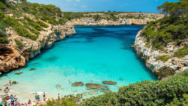Une baie côtière aux eaux turquoise et limpides, bordée de falaises rocheuses et dotée d’une plage où se trouvent des gens.