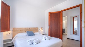 Une chambre à coucher avec une tête de lit en bois, un lit recouvert de draps blancs et agrémenté de serviettes pliées, des tables de chevet avec des lampes, et une porte en bois donnant sur une autre pièce.