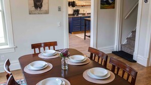 Een eetkamer met een houten tafel voor zes personen, omringd door stoelen met beklede zitplaatsen. Een kroonluchter hangt aan het plafond, en er zijn ingelijste foto's aan de muur.