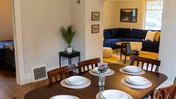 A dining room with a wooden table set for six, surrounded by chairs with patterned cushions. A chandelier hangs from the ceiling, and a potted plant is on a small table beside the dining area.
