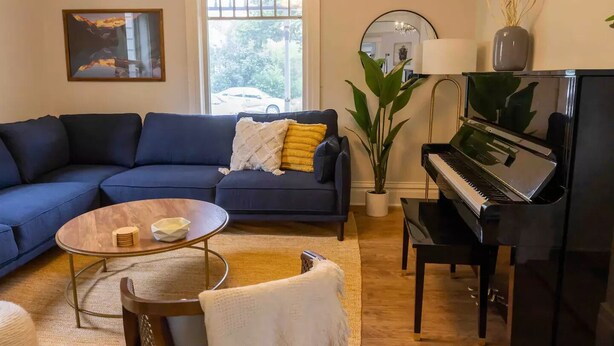 Een woonkamer met een blauwe bank, een ronde salontafel, een piano en een groot raam met uitzicht op het omliggende landschap.