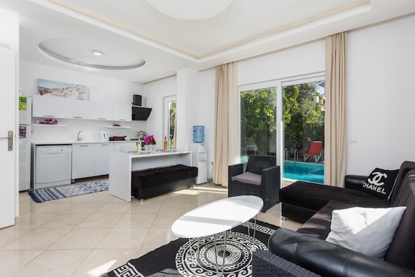 Ein modernes Wohnzimmer mit einer schwarzen Ledersofa, einem weißen Couchtisch und einem schwarz-weißen Teppich. Es gibt eine Küchenzeile mit weißen Schränken und einem Esstisch. Ein großes Fenster bietet Blick auf einen Swimmingpool und Grünflächen.