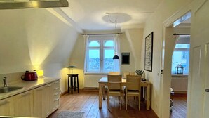 Een keuken met houten vloeren, een eetgedeelte met een tafel en stoelen, een raam met uitzicht op gebouwen, en een radiator onder het raam.