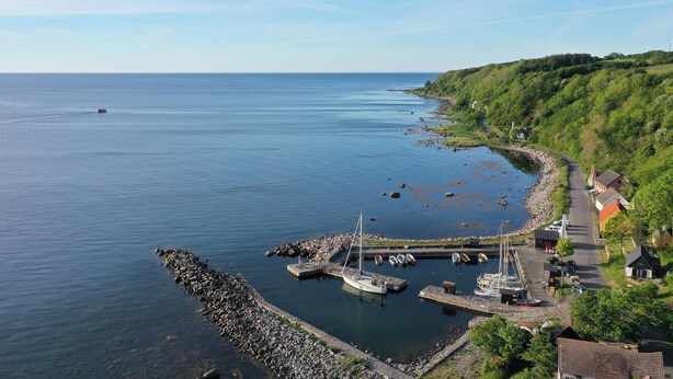 Una marina costera con barcos amarrados, un rompeolas rocoso y un camino que lleva a un pueblo con casas.