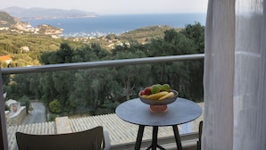 Een balkon met een tafel waarop een fruitschaal staat, met uitzicht op een kustlandschap met bomen en gebouwen.