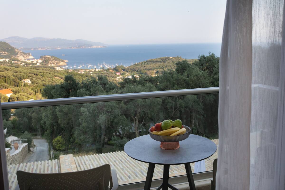 A balcony with a table holding a fruit bowl, overlooking a coastal landscape with trees and buildings.