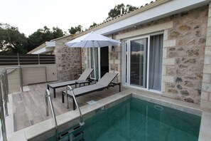 A swimming pool area with lounge chairs and an umbrella, stone walls, and a glass door. - Elais Luxury Villas (Parga)