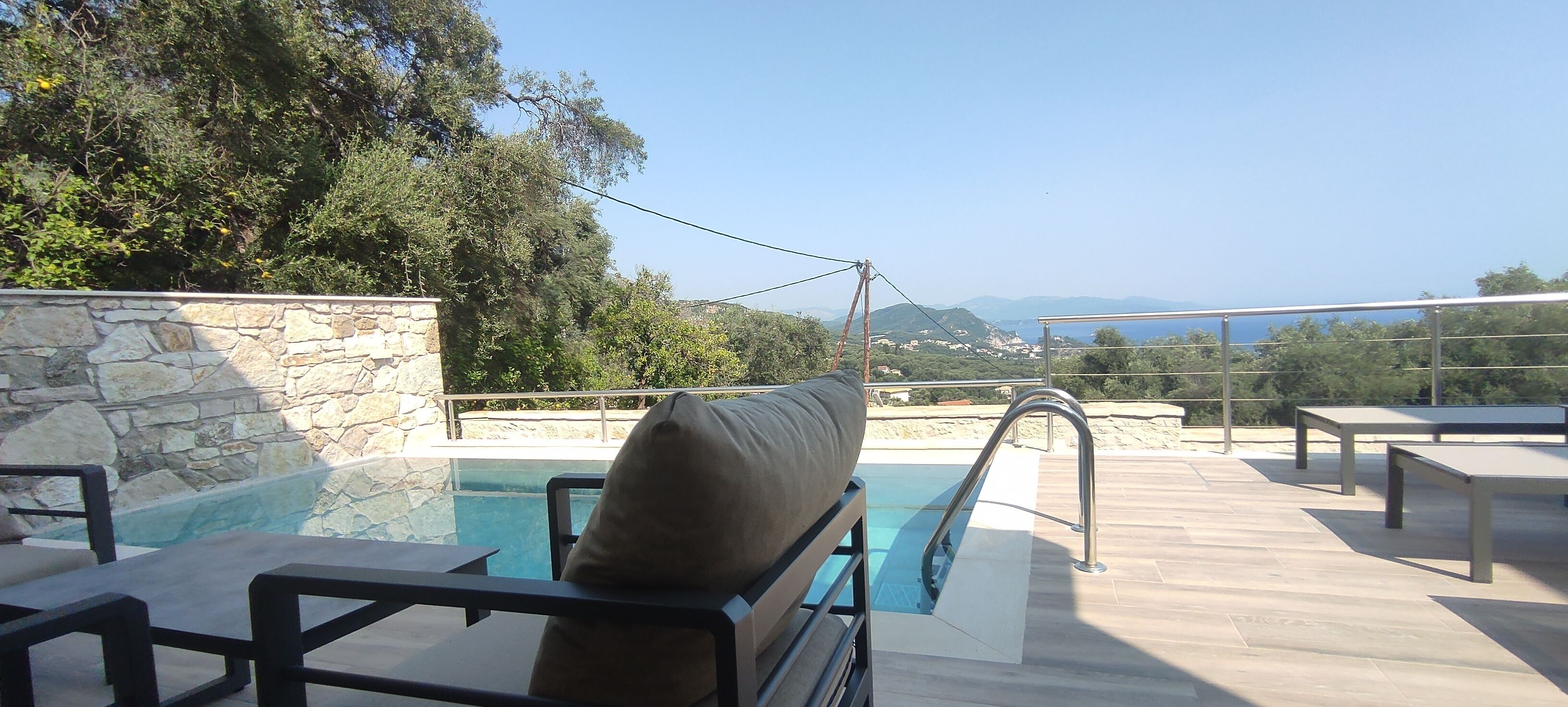A poolside area with a stone wall, lounge chairs, and a view of the sea and hills.