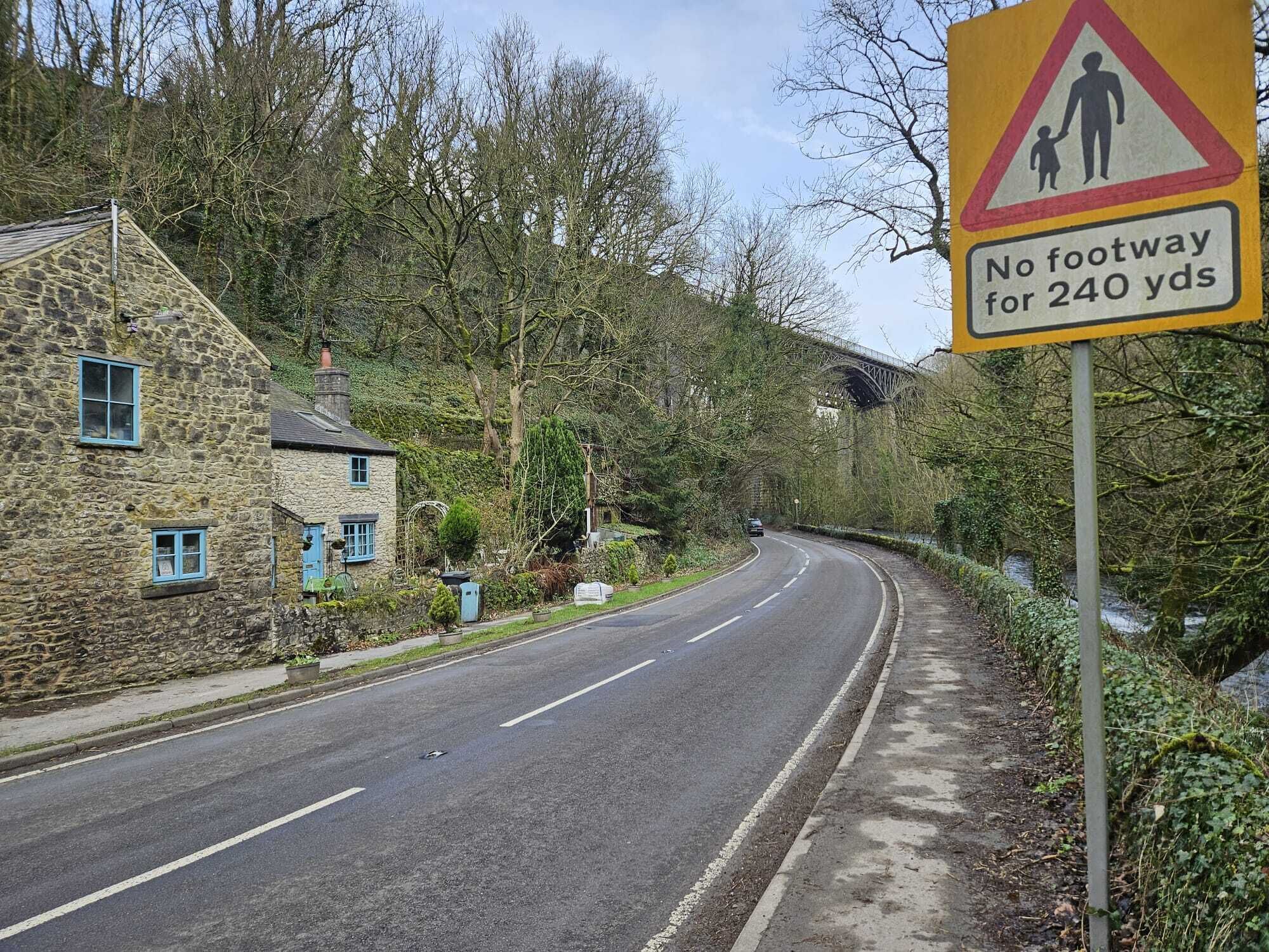 A rural road with a 'No footway for 240 yds' sign, stone houses, and a bridge in the background.