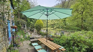 A wooden picnic table with benches and a green umbrella, surrounded by lush greenery and stone pathways. - Rose Cottage - Cosy Cottage in Millers Dale (Buxton)