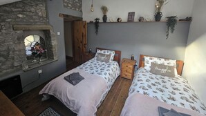 A cozy bedroom with two beds, a stone wall, a wooden ceiling, and a shelf with plants and decorative items. - Rose Cottage - Cosy Cottage in Millers Dale (Buxton)