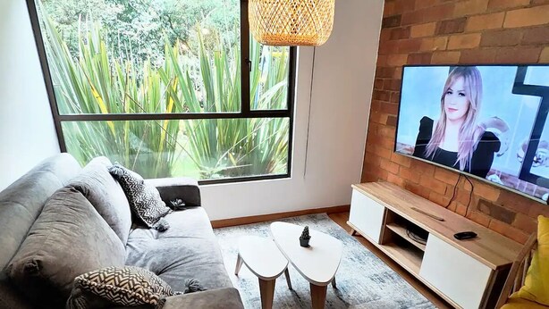 A modern living room with a grey sofa, a small white coffee table, a TV on a brick wall, and a window with a view of greenery. - Unbeatable Quality and near to the forest ! (Bogotá)