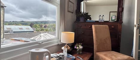 A hotel room with a wooden desk, a chair, a large window with a view of the countryside, a mirror, and a lamp.