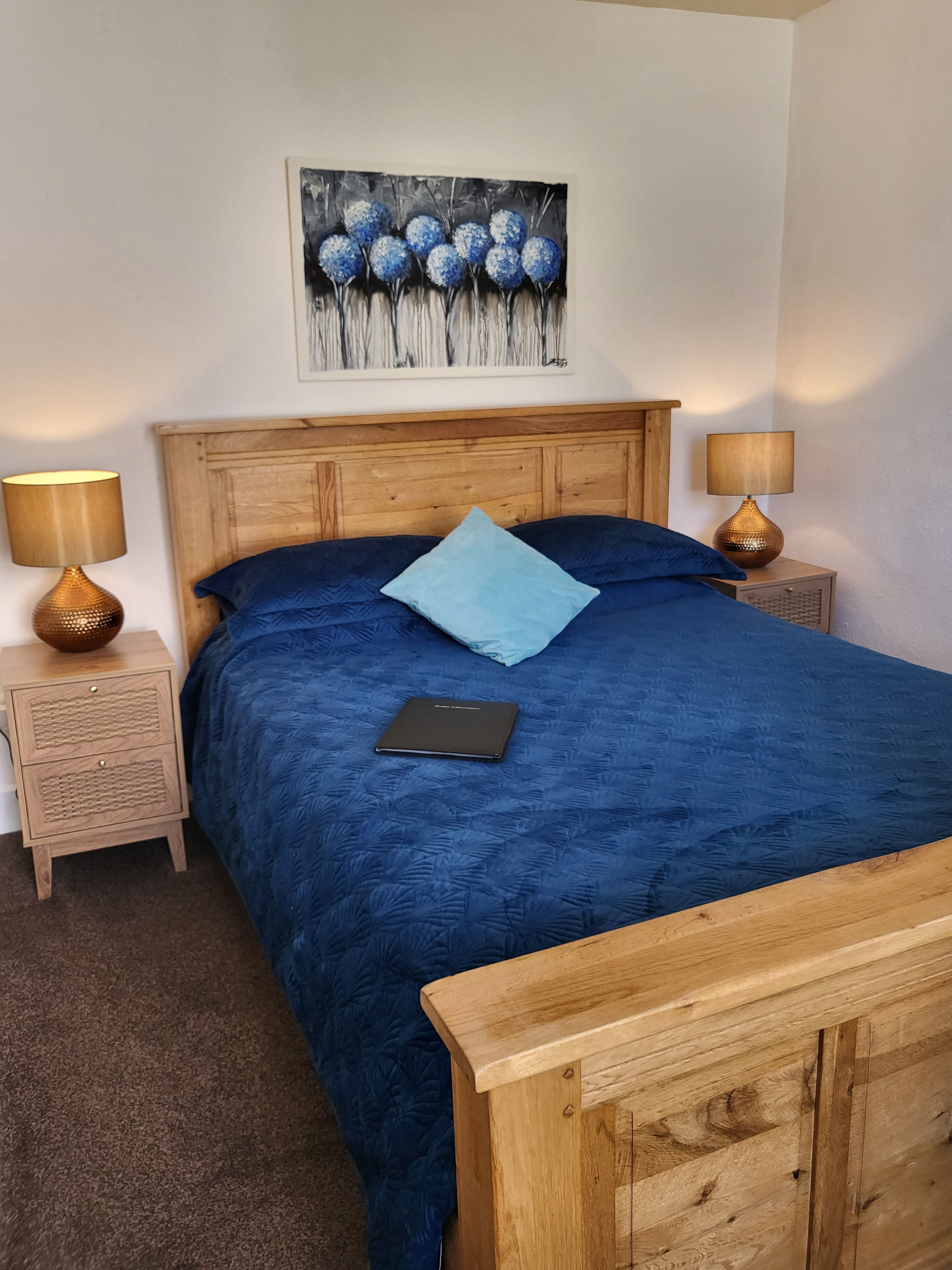 Une chambre à coucher avec une tête de lit en bois, une couvre-lit bleue, un seul oreiller, une table de chevet avec une lampe et un tableau accroché au mur.