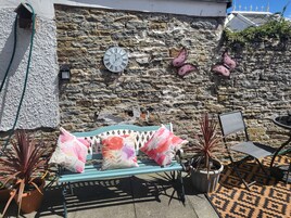 Un muro de piedra con un reloj, un banco con cojines florales y plantas en macetas.