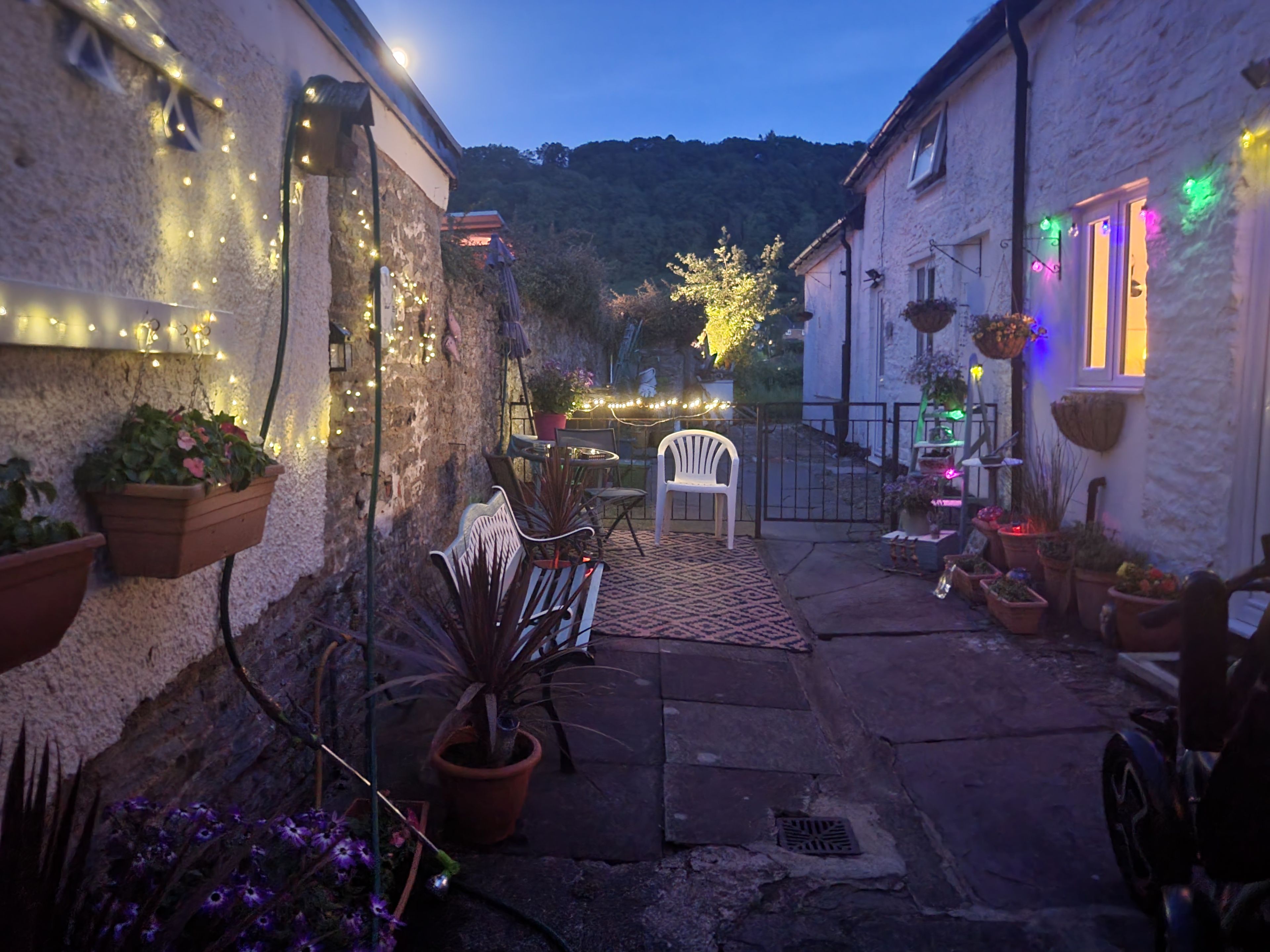Une ruelle étroite, agrémentée de plantes en pot et de quelques tables pour s’asseoir à l’extérieur, qui mène à un bâtiment illuminé de lumières colorées.
