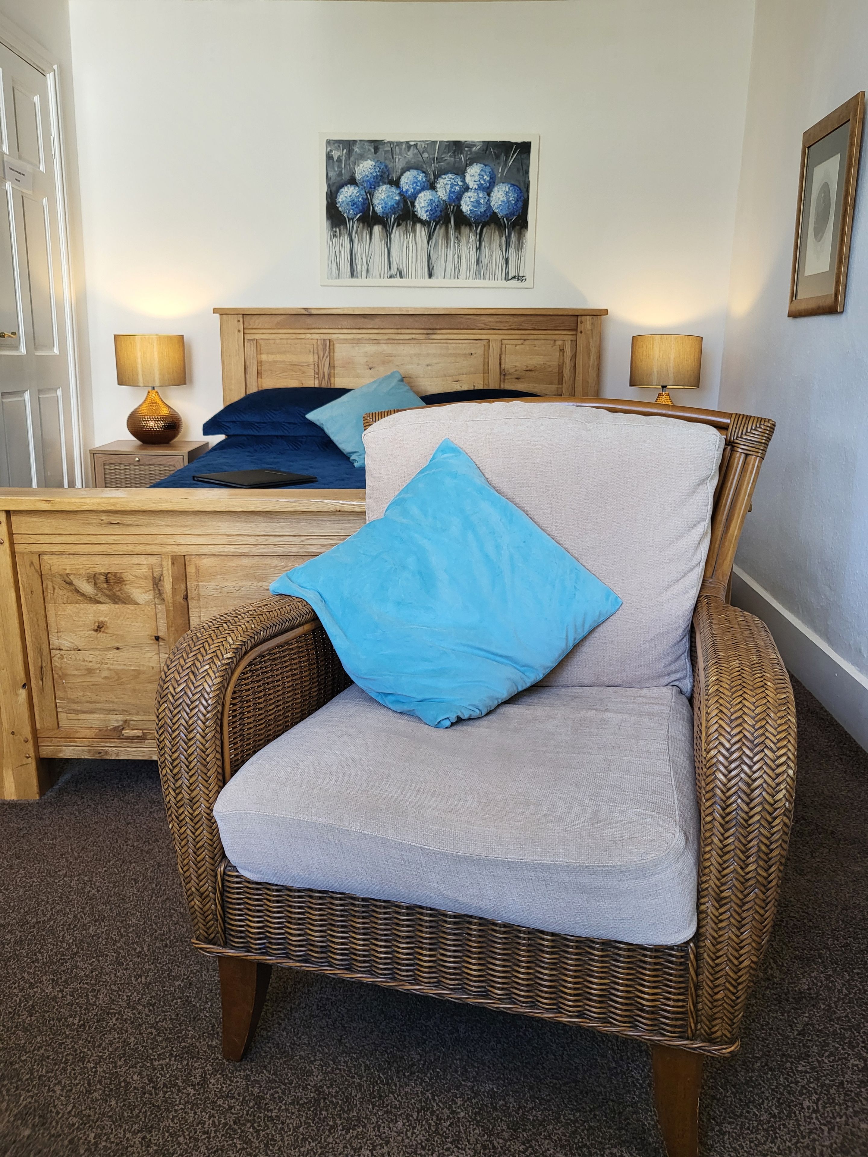 A room with a wicker chair, a bed with blue and grey bedding, a wooden headboard, and a painting on the wall.