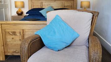 A room with a wicker chair, a bed with blue and grey bedding, a wooden headboard, and a painting on the wall.