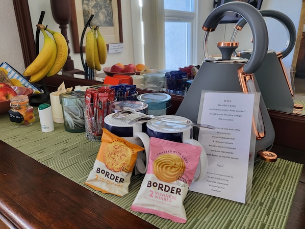 A breakfast setting with a kettle, tea bags, coffee, fruit, and packaged snacks.