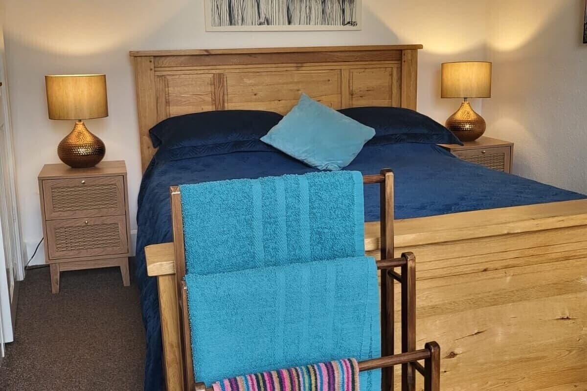 A bedroom with a wooden bed, two bedside tables with lamps, a blue blanket, and a painting on the wall.
