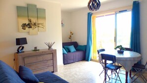 Ein Wohnzimmer mit einer Couch, einem Esstisch mit Stühlen, einem kleinen Tisch mit einer Pflanze, einer Couch mit Kissen, einer Kommode und einem Bild an der Wand.