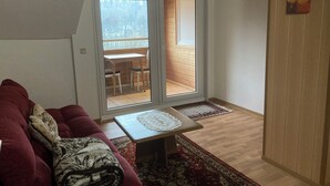 A living room with a red sofa, a small table with a lace tablecloth, and a view through a glass door to an outdoor area with a wooden deck and chairs. - Immensee I Apartment with lake view surrounded by forest (Ronshausen)