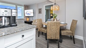 A modern kitchen with a coffee maker, dining area with wicker chairs, a television displaying 'Ocean Wave Rentals,' and a view of the ocean. - Amazing Oceanview Condo on the 17th Fl- 2 Balconies (Myrtle Beach)