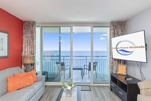 A room with a sofa, a TV, a table with a vase, and a balcony with a view of the ocean.