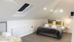Een slaapkamer met dakraam, een bed met grijze dekbedovertrek, een nachtkastje met lamp en een aan de muur bevestigde televisie.