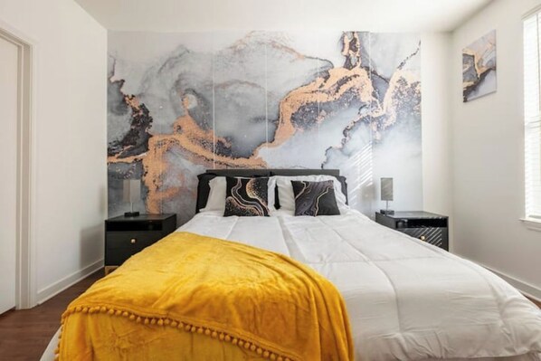A bedroom with a large bed, a yellow throw blanket, and a marble wall art piece.