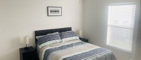 A bedroom with a bed, two bedside tables, a framed picture on the wall, and a window with blinds.