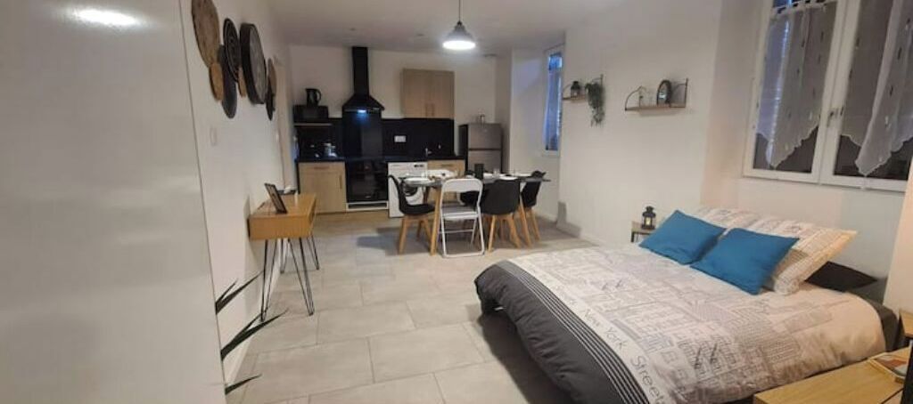 A bedroom with a bed, a desk, and a kitchenette in the background.