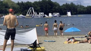 Ein Strand mit badenden und sonnenbadenden Personen, einem Rettungsschwimmturm und verschiedenen Strandutensilien.