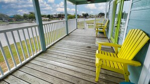 A covered wooden deck with a railing, two yellow Adirondack chairs, and a view of a body of water and distant buildings. - Creekside Getaway perfect for a relaxing weekend of fishing on the creek! (Sargent)