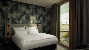Habitación de hotel moderna con una cama grande, vista de la ciudad a través de un amplio ventanal y un paisaje urbano visible.