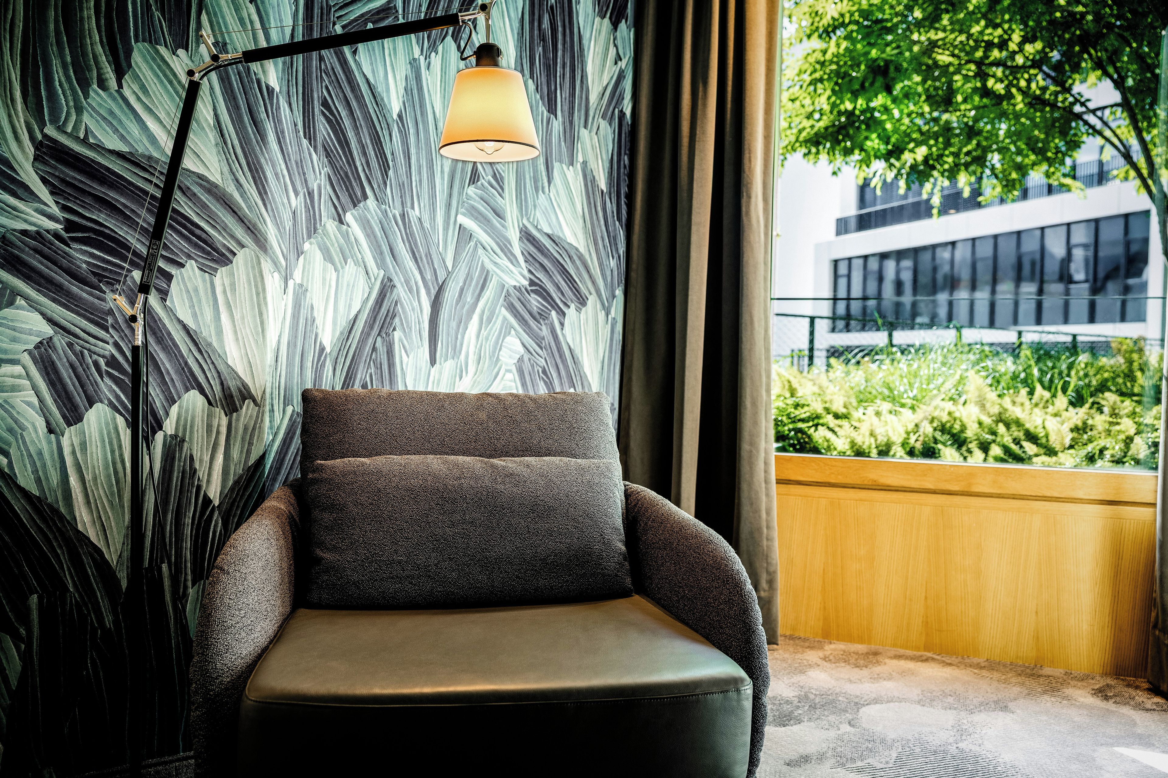 A modern armchair with a textured fabric, positioned next to a wall with a leaf pattern. A floor lamp with a curved arm is placed on the armrest.