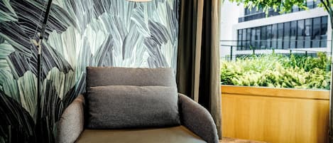 A modern armchair with a textured fabric, positioned next to a wall with a leaf pattern. A floor lamp with a curved arm is placed on the armrest.