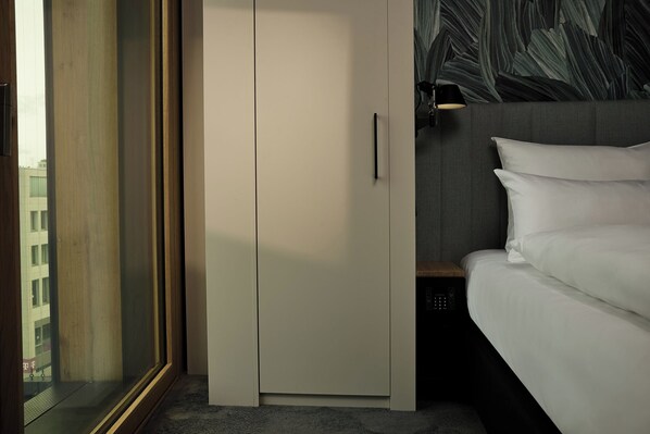 Habitación de hotel con cama, armario y vista a un paisaje urbano a través de un ventanal grande.