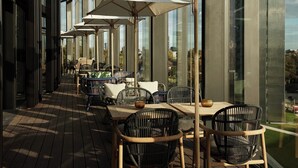 Un área de comedor al aire libre con mesas y sillas, grandes sombrillas y vistas de la ciudad a través de amplios ventanales.