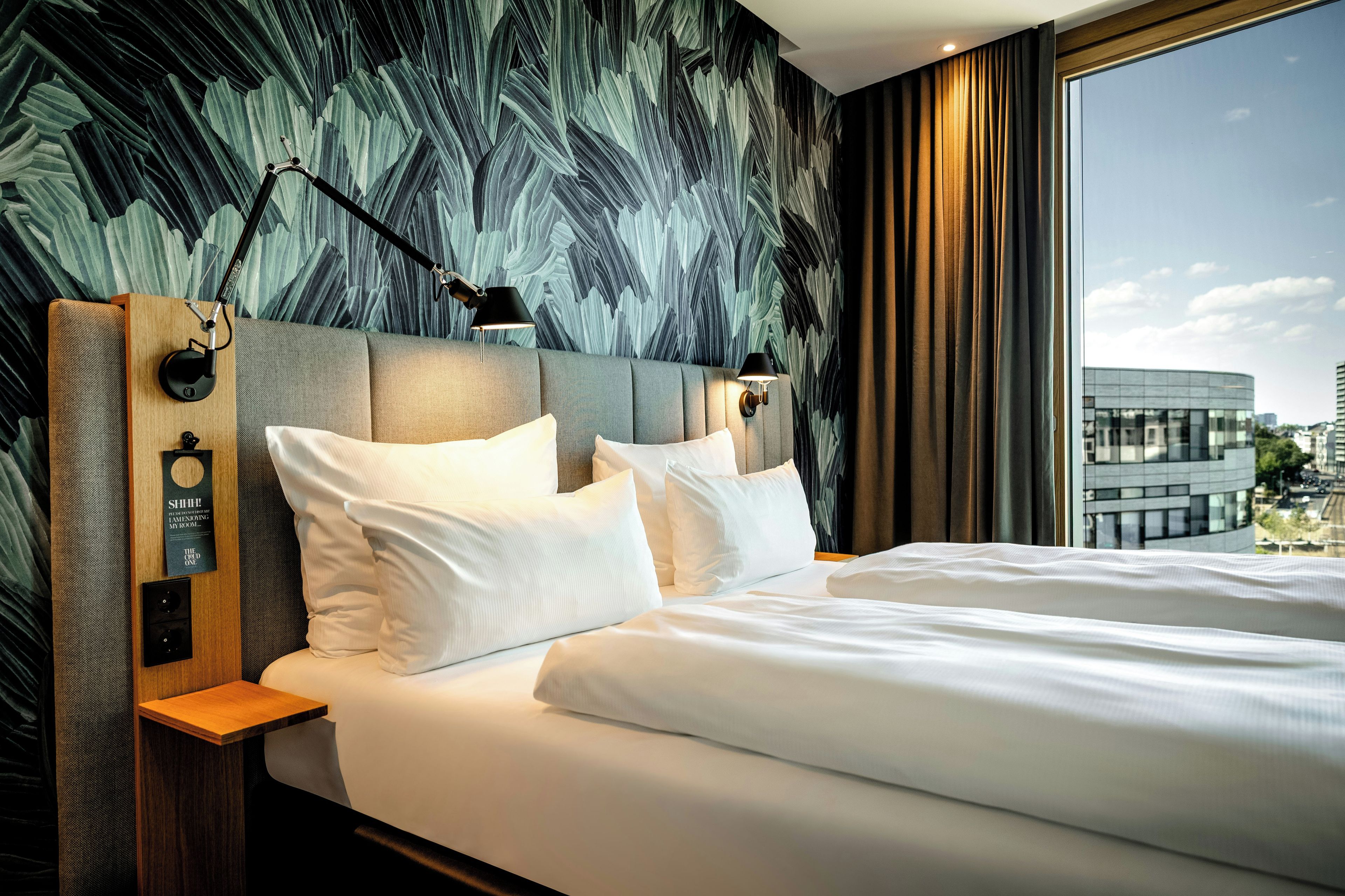 Habitación de hotel con una cama grande, dos lámparas de noche, una mesita auxiliar y vistas a la ciudad a través de una ventana.