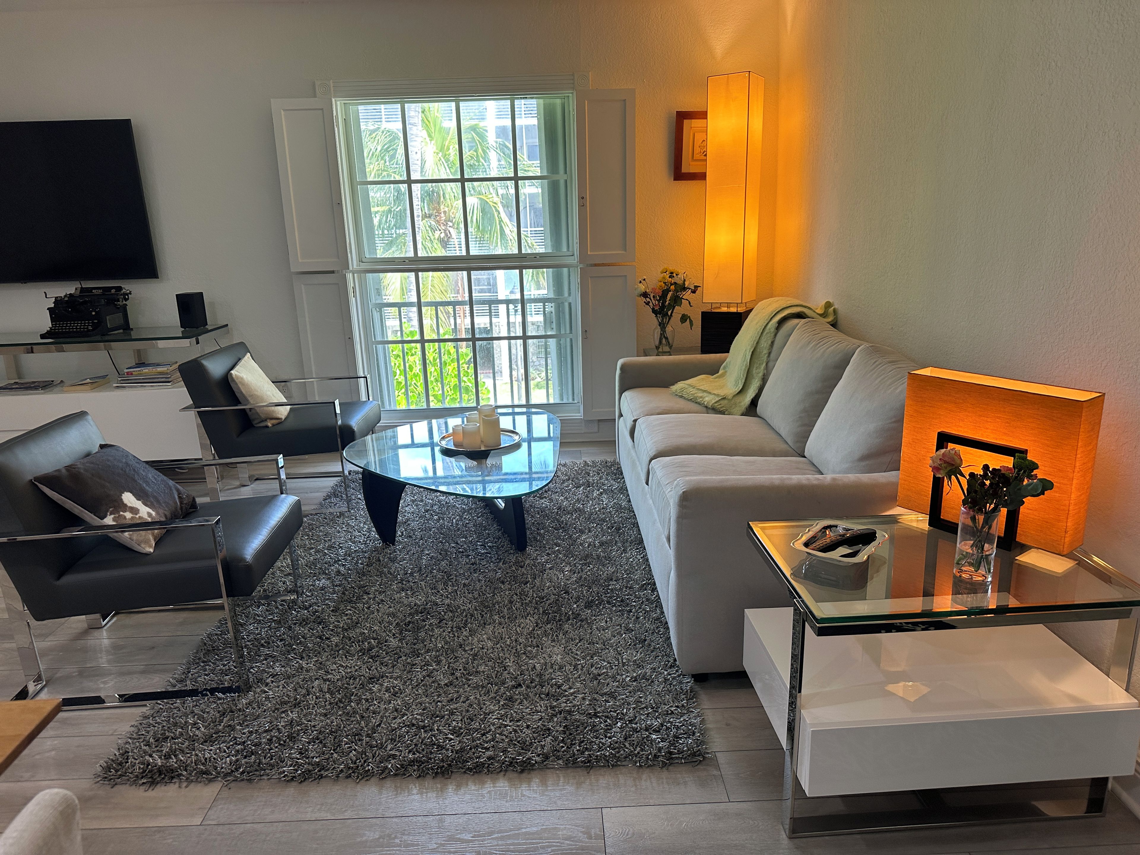 A modern living room with a sofa, armchairs, a glass coffee table, and a TV on a wall.