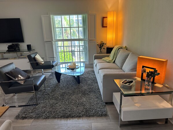 A modern living room with a sofa, armchairs, a glass coffee table, and a TV on a wall.