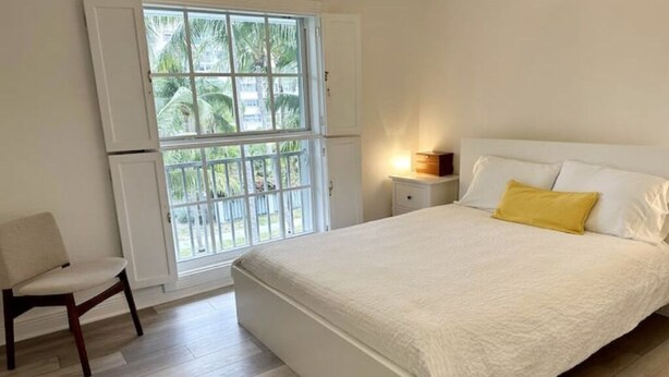 Ein Schlafzimmer mit Bett, Stuhl, Nachttisch und einem Fenster mit Blick auf Palmen.
