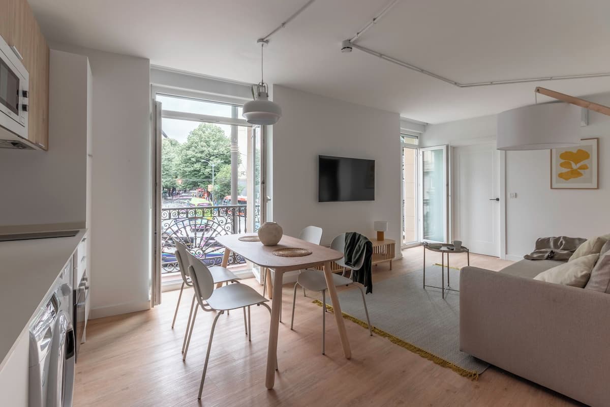 A modern living room with a sofa, a dining table with chairs, a TV mounted on the wall, and a balcony with a view of the outside.