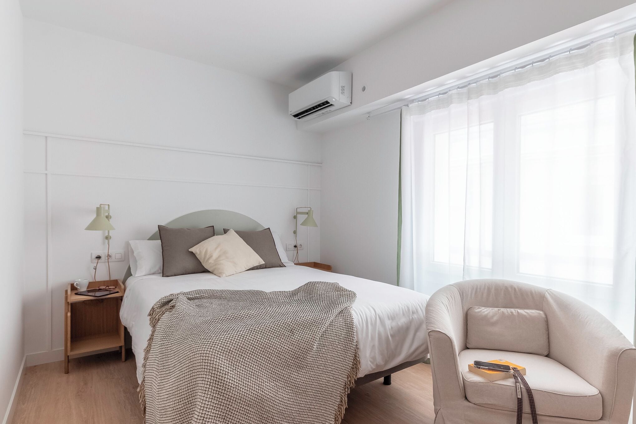 A modern hotel room with a neatly made bed, a bedside table with a lamp, a chair, and a large window with sheer curtains.